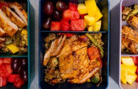 A high-protein meal prep container featuring sliced sriracha chicken over a bed of quinoa with roasted vegetables and a side of fresh fruit.