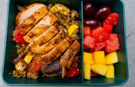 A high-protein meal prep container featuring sliced sriracha chicken over a bed of quinoa with roasted vegetables and a side of fresh fruit.