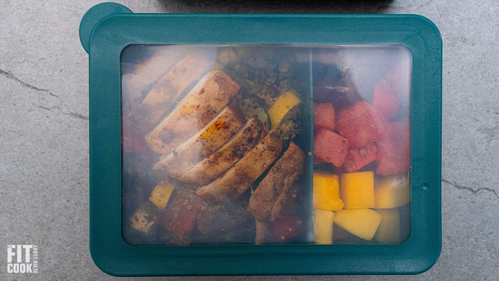 A high-protein meal prep container featuring sliced sriracha chicken over a bed of quinoa with roasted vegetables and a side of fresh fruit."