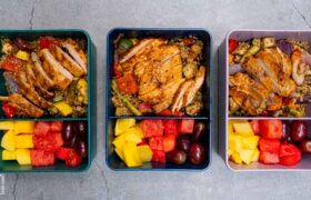 A high-protein meal prep container featuring sliced sriracha chicken over a bed of quinoa with roasted vegetables and a side of fresh fruit.