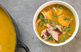 Bowl of Caribbean seafood soup topped with flaked salmon and scallion greens next to the pot, a meal prep protein topper