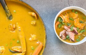 Overhead of a pot of Caribbean seafood soup with crab legs beside a finished bowl topped with flaked salmon and scallions