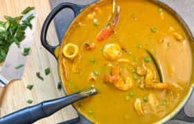 Caribbean seafood soup in a cast iron pot next to a cutting board of chopped scallions, with shrimp and scallops in a sweet potato coconut broth